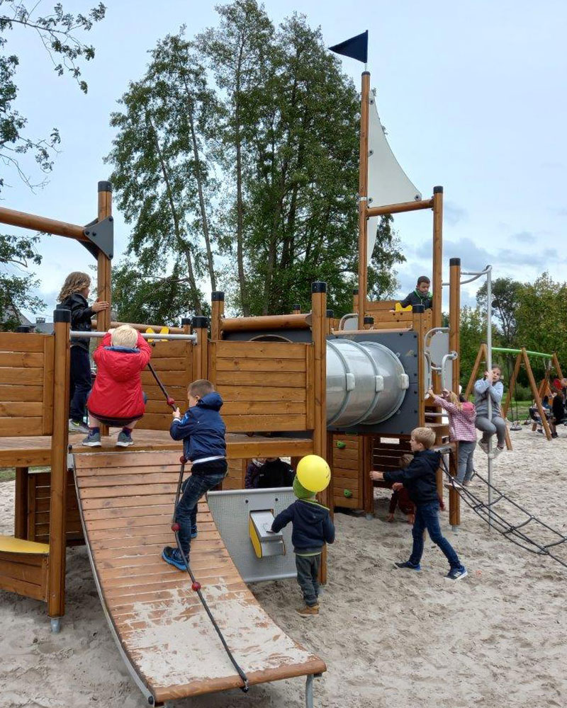 I bambini salgono a bordo di un sistema di gioco UniPlay a forma di barca, realizzato in legno. È situato su una superficie sabbiosa.